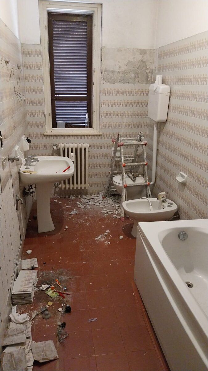 Bagno prima dell'intervento: sanitari e radiatore originali, rivestimento datato, macerie a terra.